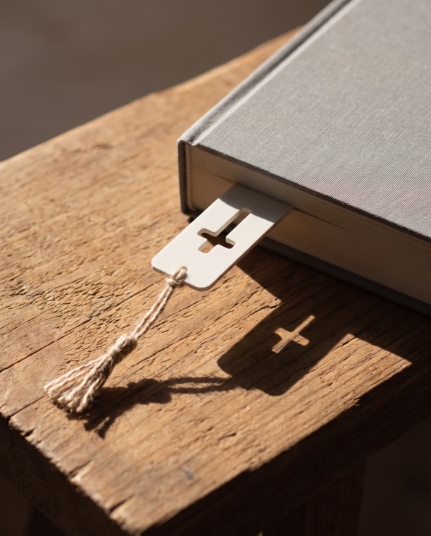 Marca-página em forma de cruz, resina cor cal, dentro de um livro sobre madeira crua.